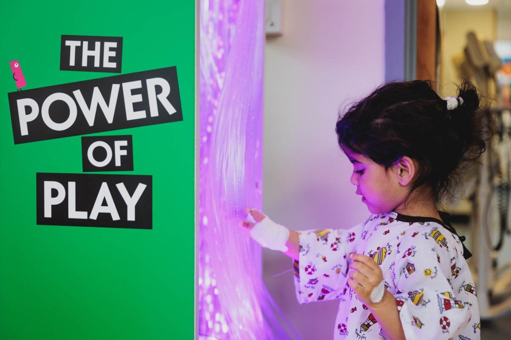 Young girl in hospital gown is mesmerised by sparkling lights on a sensory unit. A sign reading "The Power of Play" is in the background.